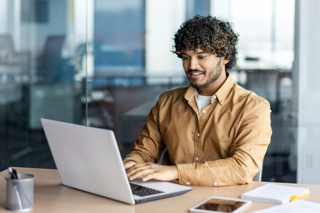 Feliz y sonriente hombre de negocios hispano escribiendo en una computadora portátil, oficinista con cabello rizado feliz con