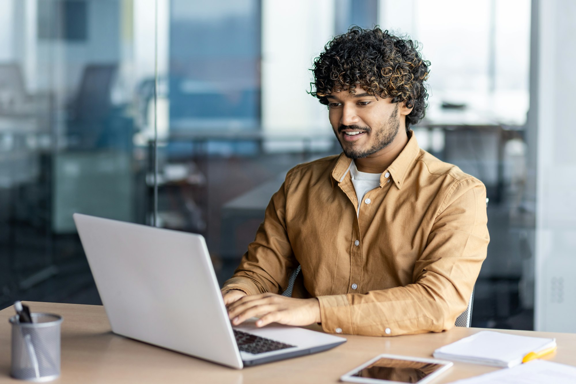 Feliz y sonriente hombre de negocios hispano escribiendo en una computadora portátil, oficinista con cabello rizado feliz con