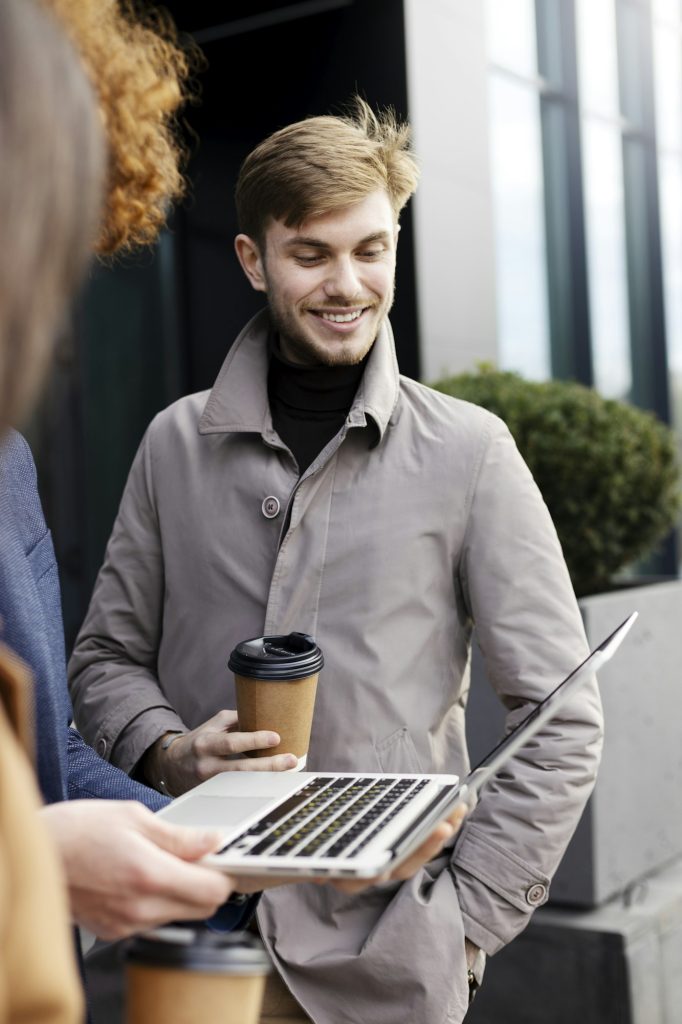 Successful man standing outdoors, talking to his co-worker, looking at laptop.